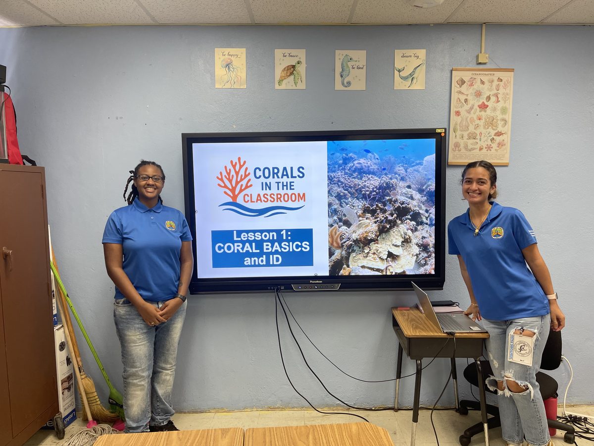 Two Reef Response staff members beside a classroom screen showing the Corals in the Classroom title slide for Lesson 1, Coral Basics and ID